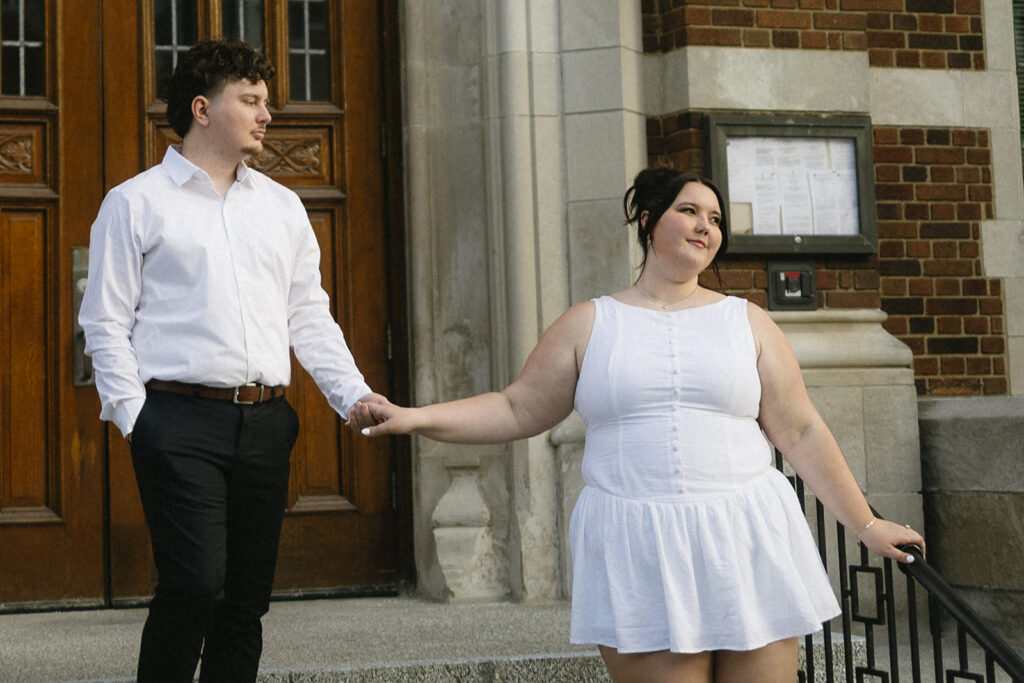 A couple in white clothes holds hands on stone steps outside a building with wooden doors and brick walls. The woman stands in front, looking away, while the man looks at her.