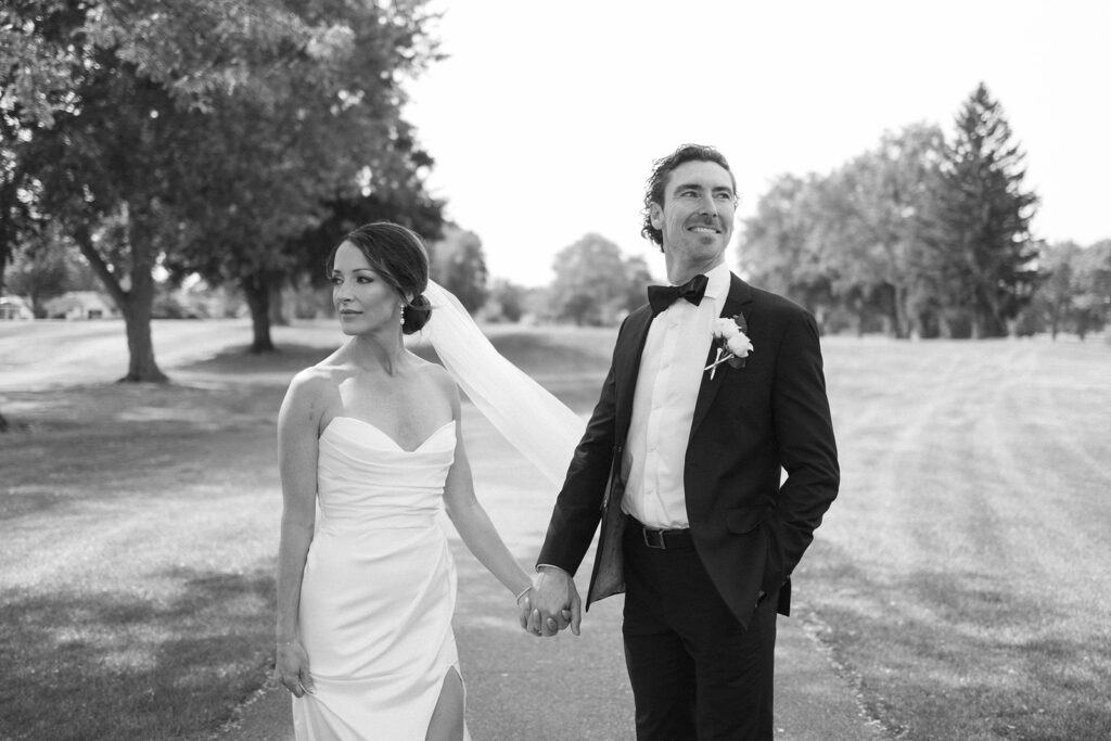 A bride in a strapless gown and a groom in a tuxedo with a bow tie hold hands outdoors. The bride looks left while the groom looks right, both standing on a tree-lined path. The image is black and white.