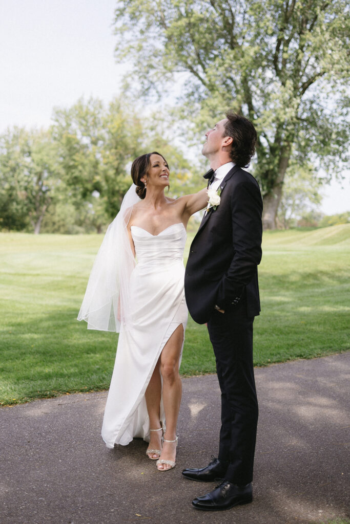 A bride in a white dress and veil smiles up at a groom in a black tuxedo as they stand on a path in a green park with trees in the background. The groom is looking up, and they seem joyful and relaxed.