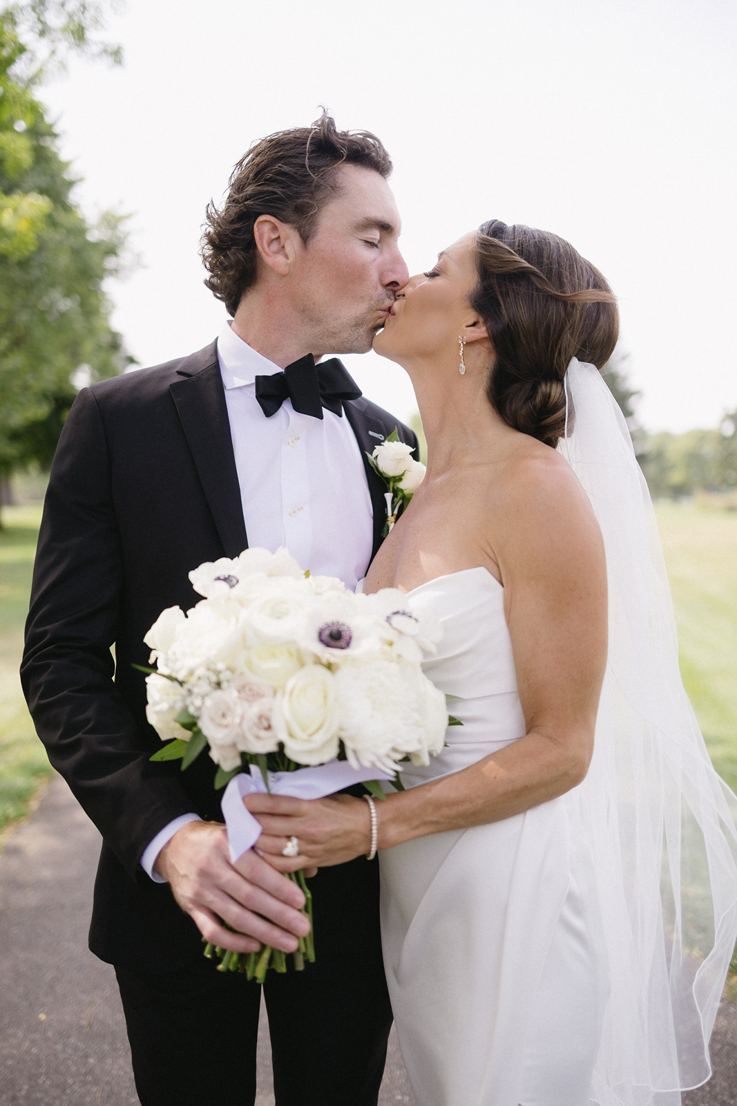 A bride and groom in formal wedding attire share a kiss outdoors. The bride holds a bouquet of white flowers and wears a strapless dress and veil; the groom wears a black tuxedo with a white shirt and bow tie.