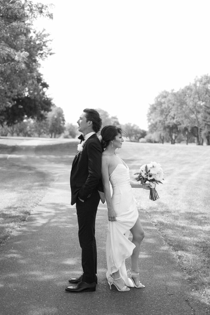 A bride and groom stand back-to-back on a tree-lined path. The bride holds a bouquet and lifts her dress slightly, while the groom stands with hands in pockets. Both are smiling. The scene is lit by soft daylight.