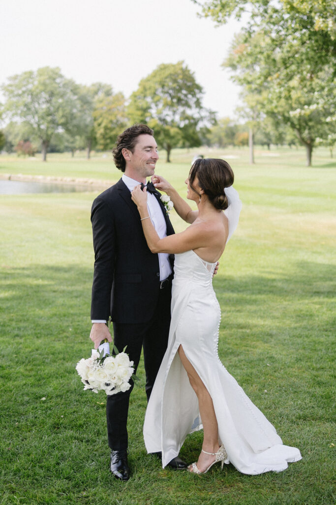 A bride in a white dress adjusts the groom’s bowtie as they stand on grass outdoors. The groom holds a bouquet of white flowers; both are smiling amid green trees and bright daylight.