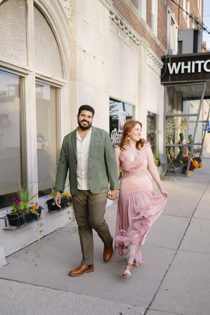 A couple holds hands while walking on a city sidewalk; the man wears a green blazer and khaki pants, and the woman wears a flowing pink dress, looking at him and smiling. They pass flowers and a building with a WHITC sign.