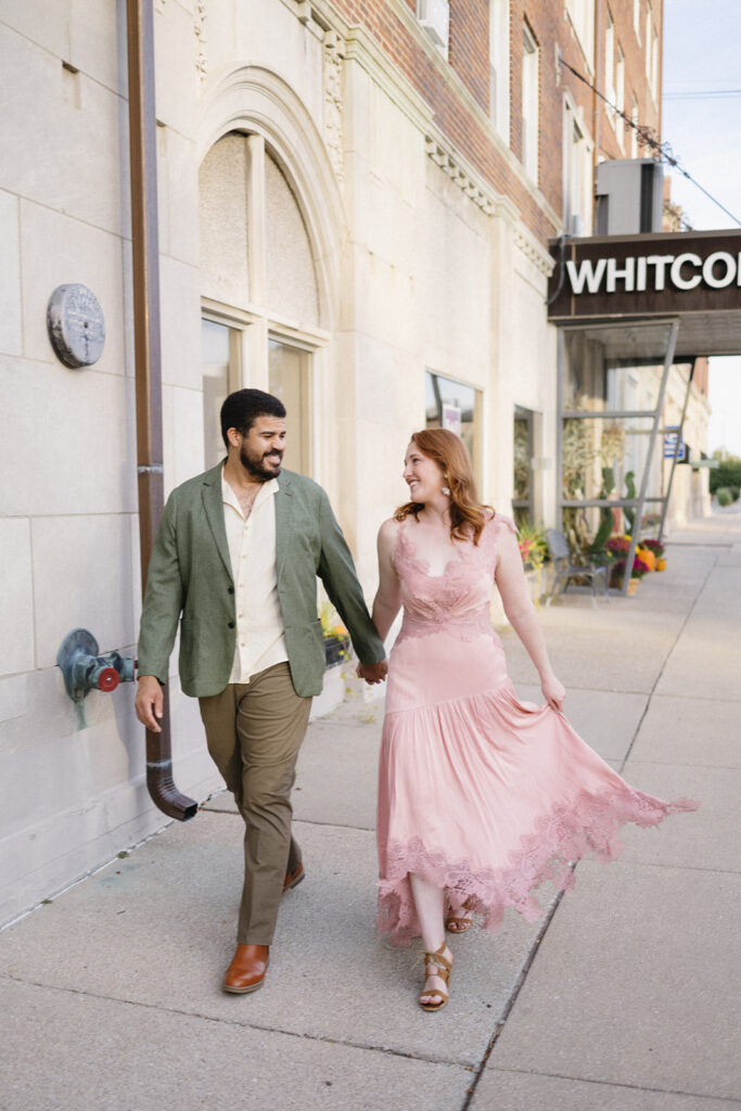 A couple holding hands walks down a city sidewalk, smiling at each other. The man wears a green blazer and khaki pants; the woman wears a pink dress. They pass by a building with a WHITCOM sign.