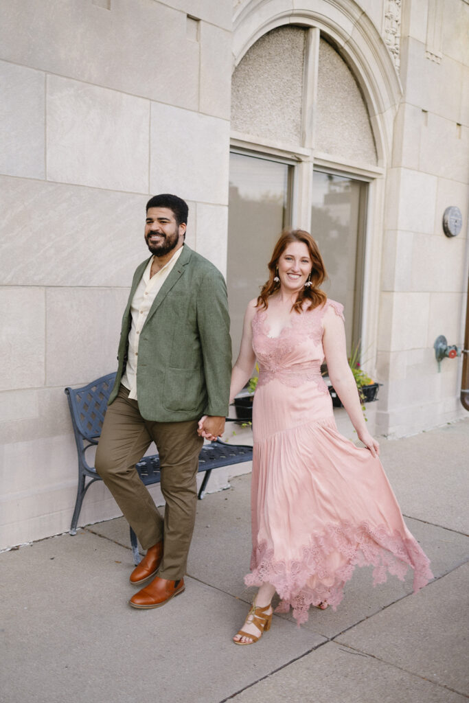 A smiling couple holds hands while walking outside. The man wears a green blazer, cream shirt, and brown pants. The woman wears a flowing pink dress and gold sandals. Theyre on a sidewalk near a stone building with arched windows.
