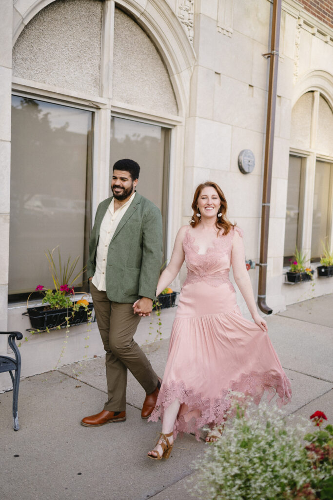 A smiling couple walks hand-in-hand on a sidewalk. The man wears a green blazer and khaki pants; the woman wears a flowing pink dress and sandals. They pass by a building with arched windows and flower boxes.