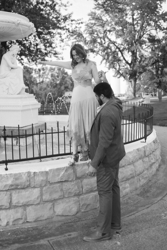 A man in a suit helps a woman in a dress balance on the edge of a stone fountain in a park. The scene is black and white, with trees and statues visible in the background.