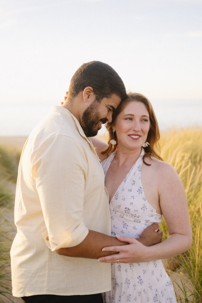 A couple embraces and smiles while standing in tall grass by the beach, bathed in warm sunlight. The woman wears a light floral dress, and the man wears a light-colored shirt.
