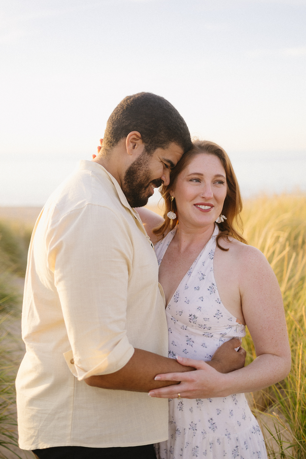 A couple embraces and smiles while standing in tall grass by the beach, bathed in warm sunlight. The woman wears a light floral dress, and the man wears a light-colored shirt.