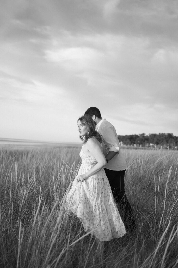 A woman in a light dress and a man stand close together in tall beach grass, with the shoreline and trees in the background under a partly cloudy sky. The image is in black and white.