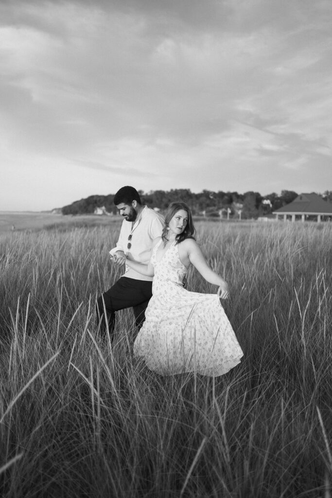 A woman in a flowing dress and a man walk through tall grass in an open field near the coast, with houses and trees in the background under a partly cloudy sky. The image is in black and white.