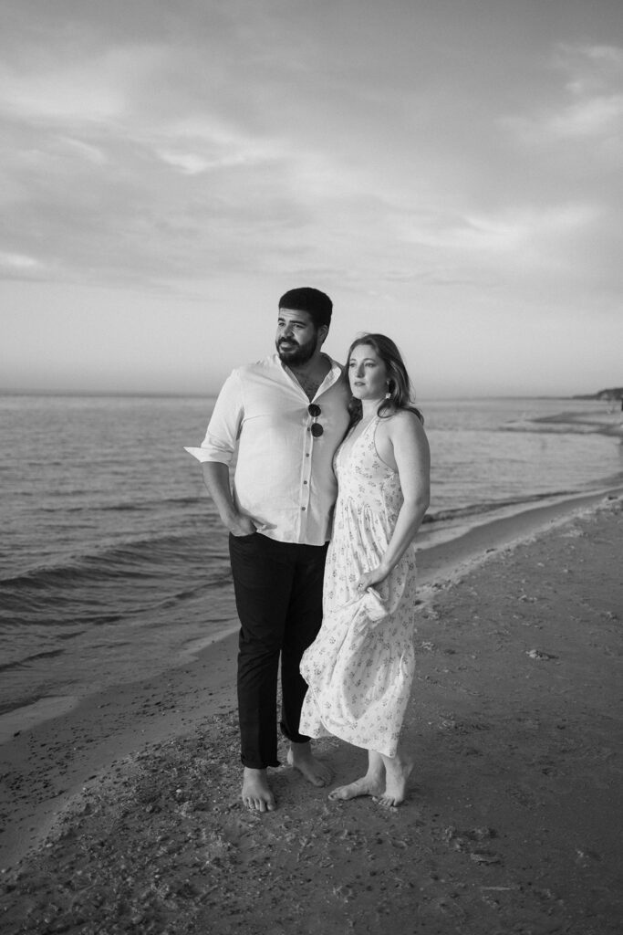 A man and woman stand barefoot on a beach at sunset, looking off into the distance. The man wears a white shirt and dark pants; the woman wears a light, sleeveless dress. The scene is calm and romantic, in black and white.