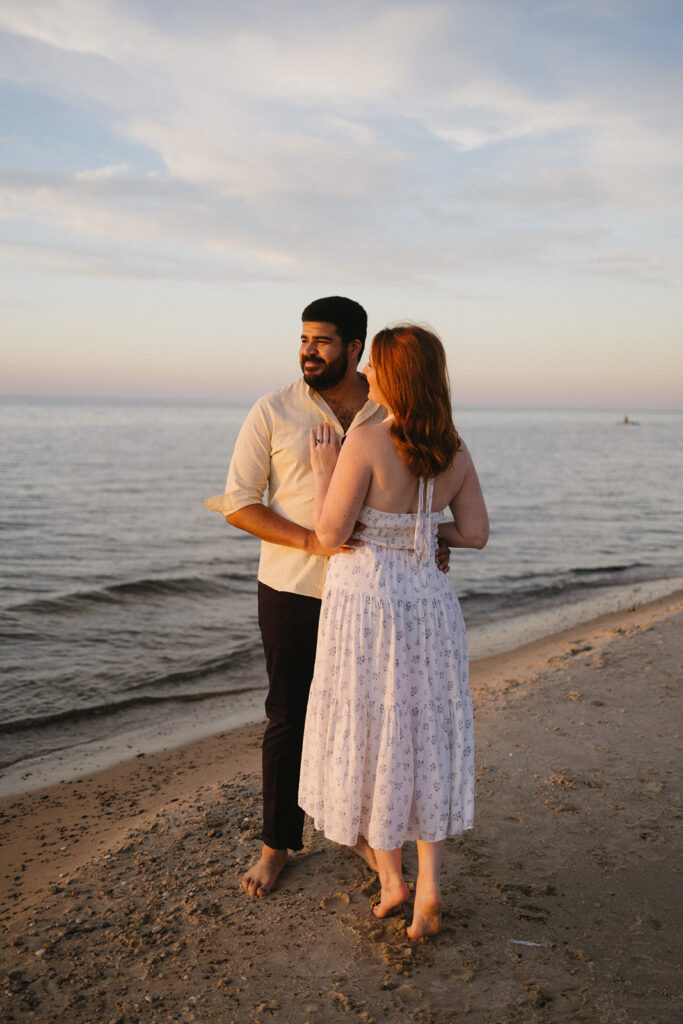 A couple stands barefoot on a sandy beach at sunset, embracing and looking into the distance. The woman wears a white summer dress and the man wears a light shirt and dark pants. Calm waves and a pastel sky are in the background.