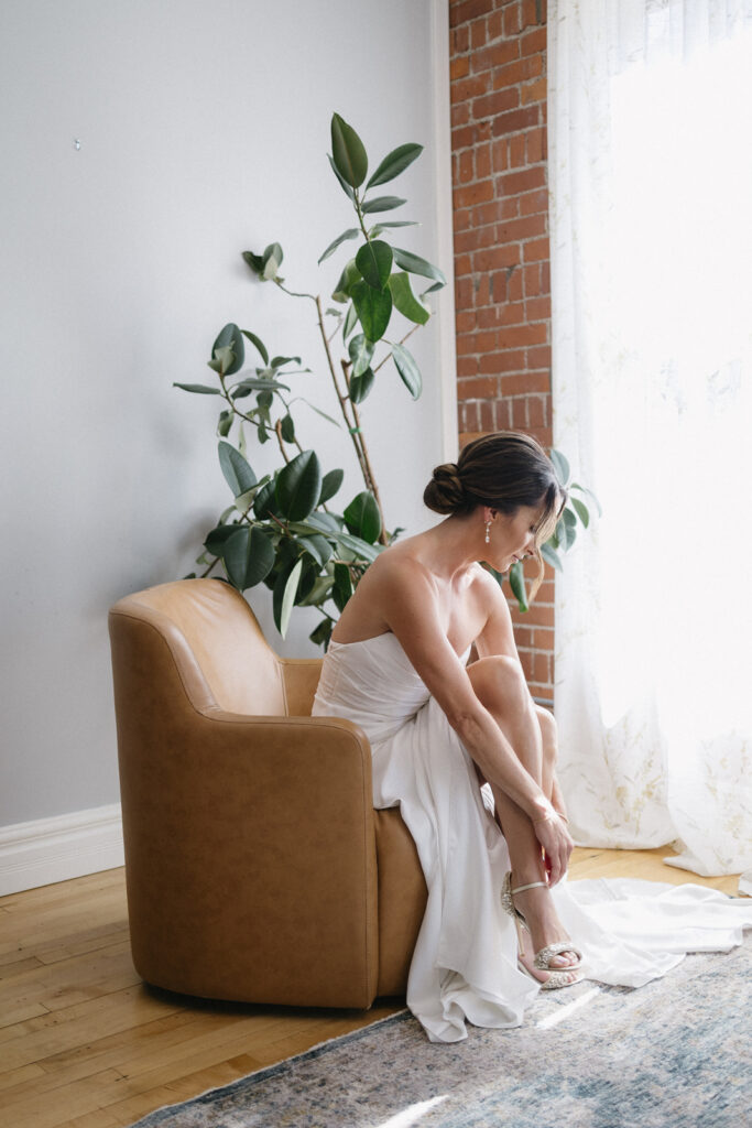 A bride in a strapless white wedding dress sits on a tan armchair near a window with sheer curtains, adjusting her sandal. A large potted plant and exposed brick wall are in the background.