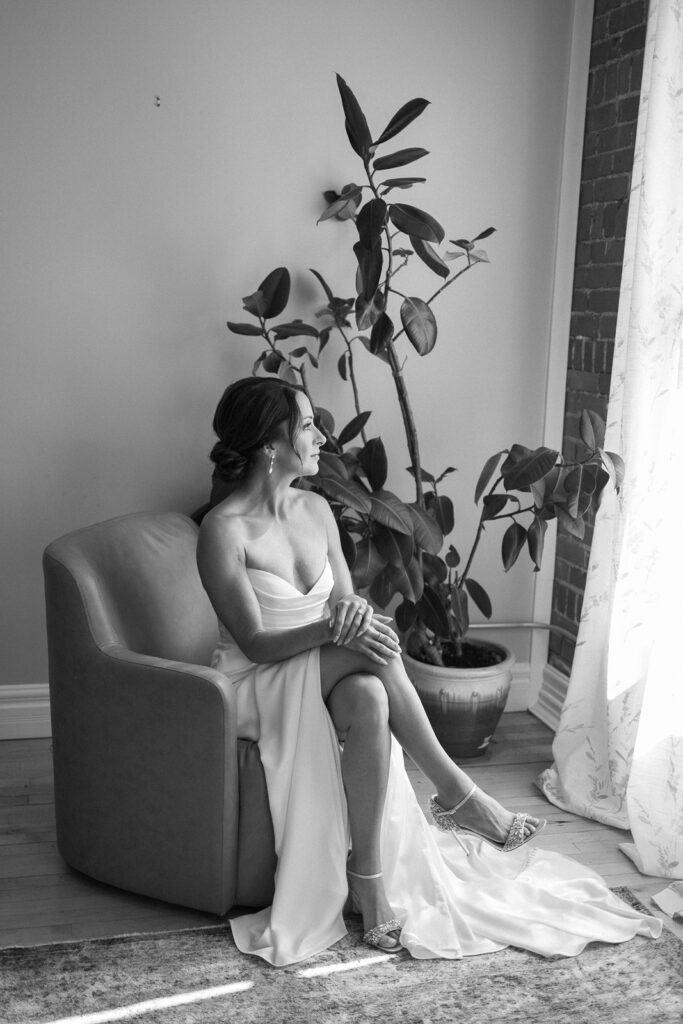 A bride in a strapless gown sits in an armchair by a window, looking outside. She is barefoot, with one leg crossed over the other. A large potted plant and brick wall are in the background. The image is in black and white.