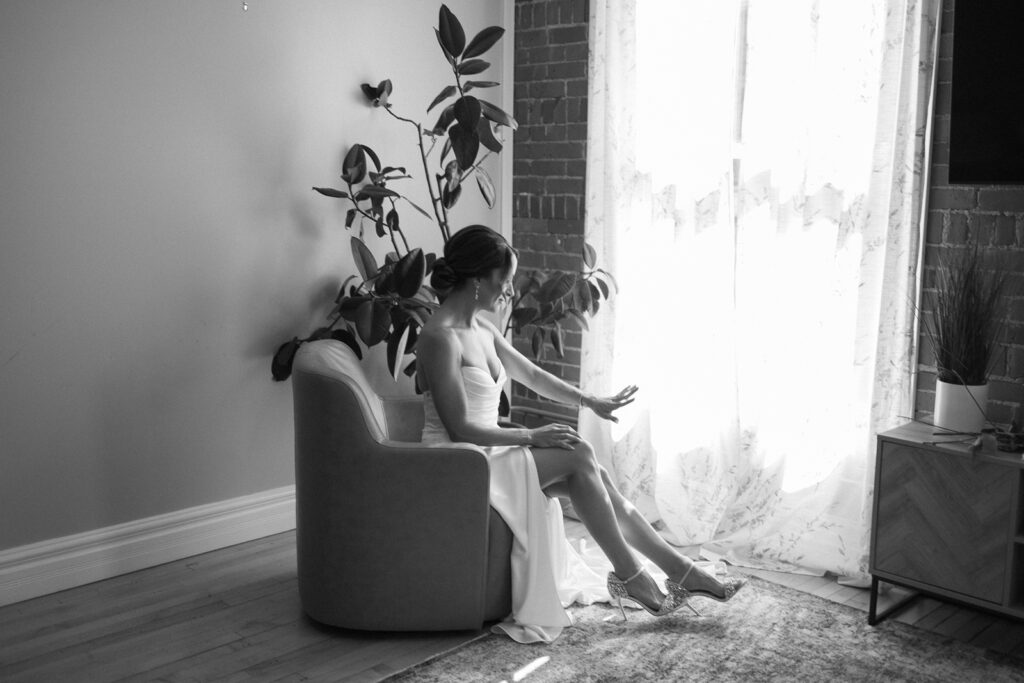 A woman in a dress sits in an armchair by a window with sheer curtains, reaching toward the sunlight. A potted plant and modern decor surround her. The scene is in black and white.