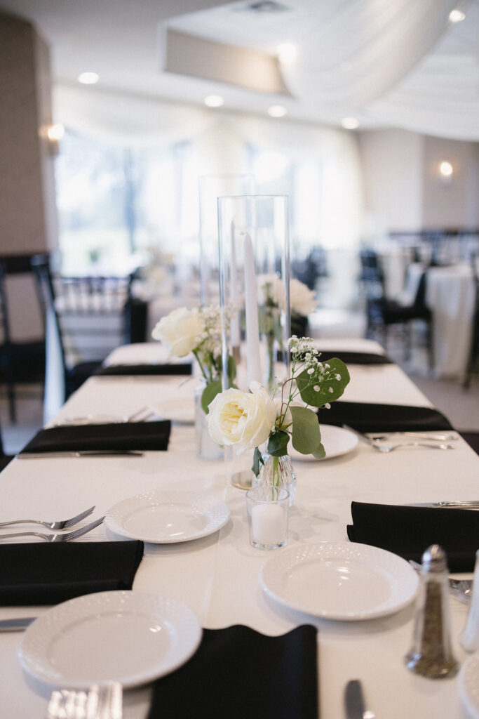 A formal table setting with white plates, black napkins, silver cutlery, and small flower arrangements featuring white roses and greenery on a white tablecloth in an elegant, softly lit event space.