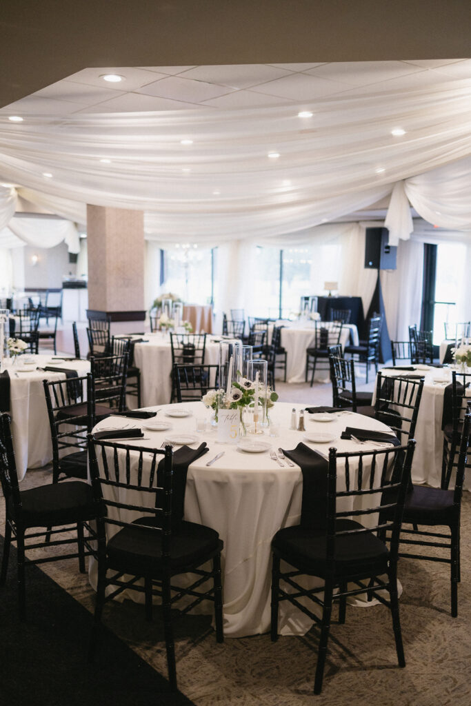 A banquet hall set for an event, featuring round tables with white tablecloths, black chairs, and simple floral centerpieces. The ceiling is draped with white fabric, creating an elegant atmosphere.