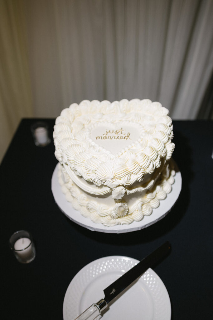 A heart-shaped white cake with piped icing and just married written on top sits on a white stand. A plate, knife, and candles are nearby on a black tablecloth.