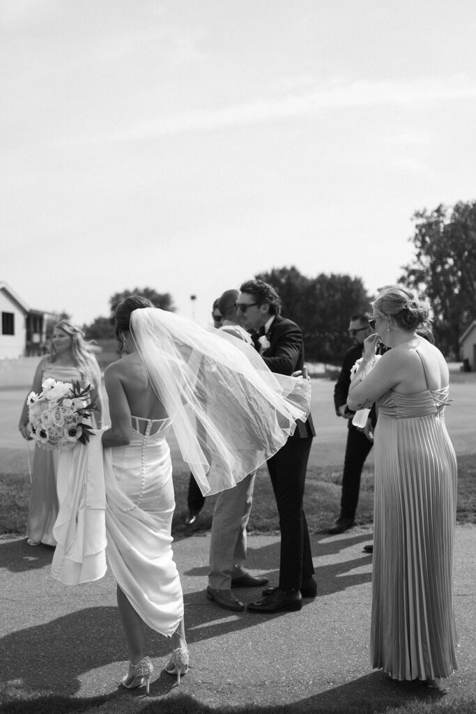 A bride in a flowing dress and veil stands outdoors with bridesmaids and groomsmen. The group is casually gathered on a sunny day, with bouquets and formal attire visible. The scene is candid and joyful.