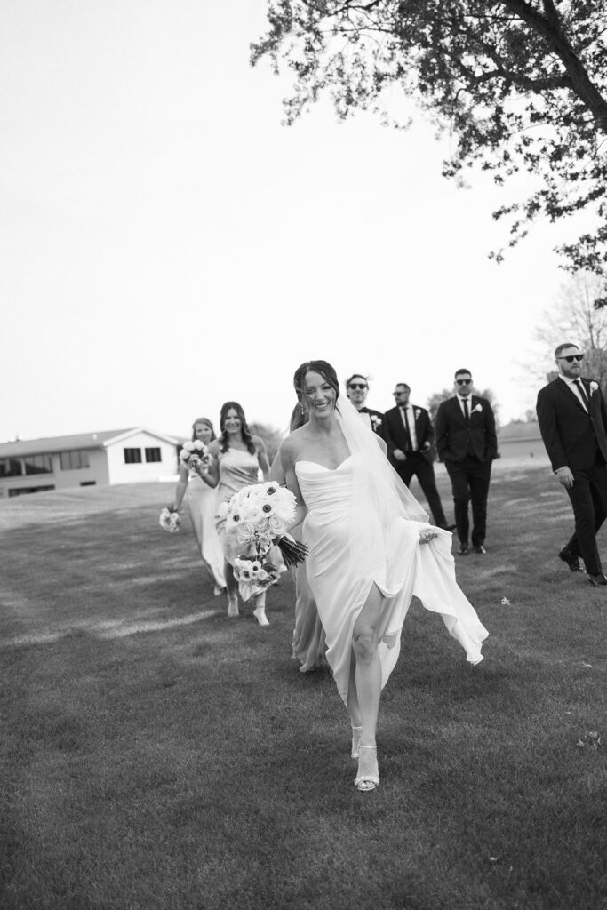 A bride in a flowing dress walks across a grassy lawn, smiling and holding her bouquet, followed by bridesmaids and groomsmen in formal attire on a sunny day.