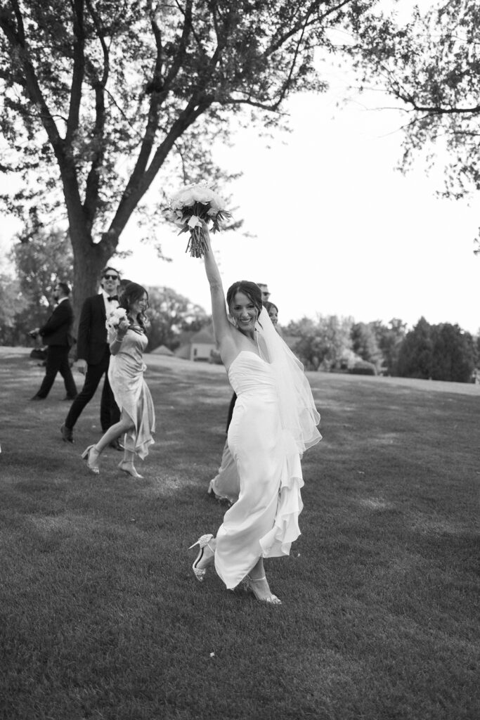 A joyful bride in a flowing white dress holds her bouquet high and smiles while walking barefoot on grass, with wedding guests in the background under tall trees.