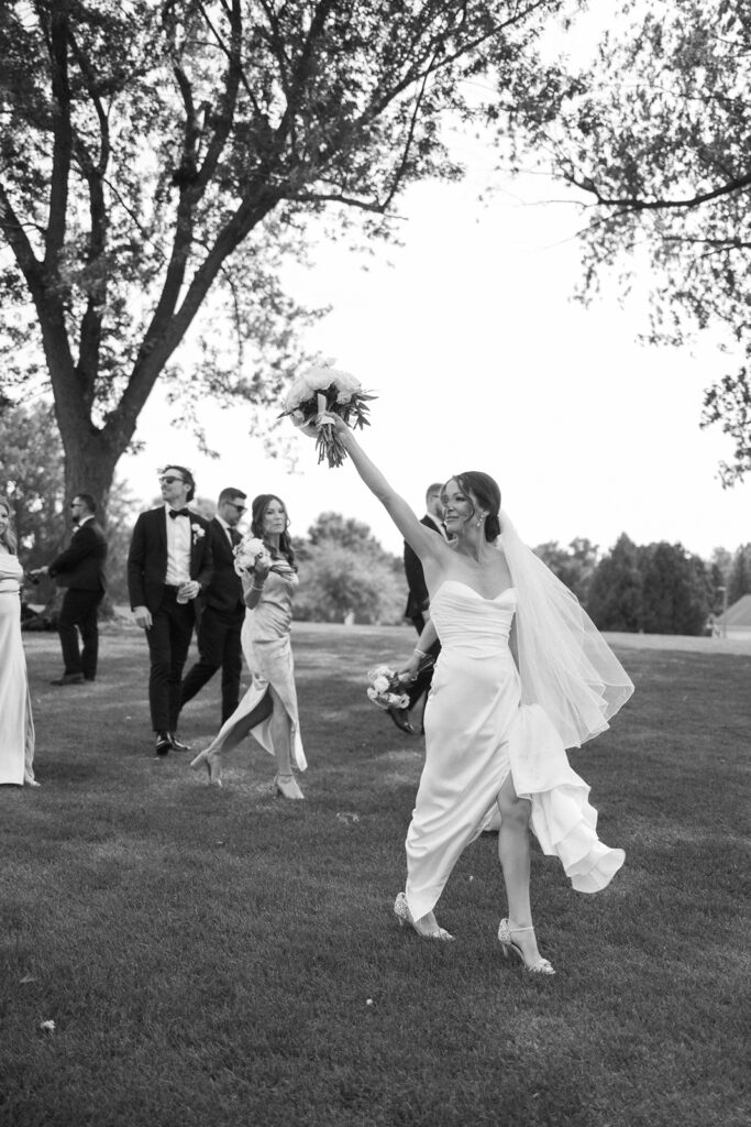 A bride in a wedding gown and veil walks on grass, holding a bouquet aloft and smiling, followed by bridesmaids and groomsmen dressed formally. Trees and open lawn appear in the background.