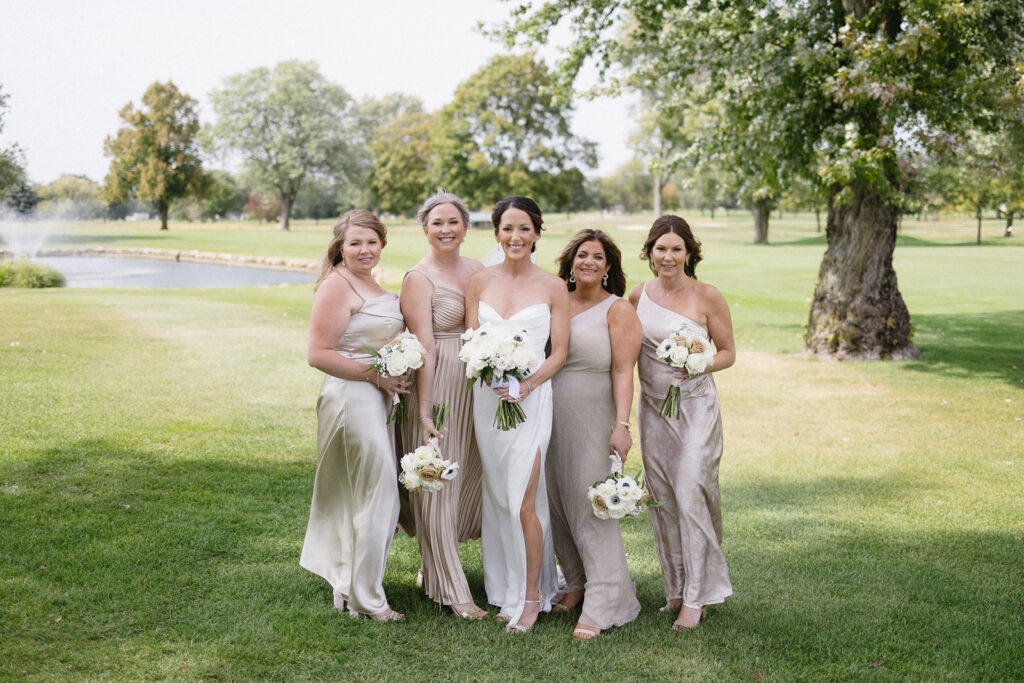 Five women in light-colored dresses stand together on grass, holding white bouquets and smiling at the camera, with trees and a small lake in the background on a sunny day.