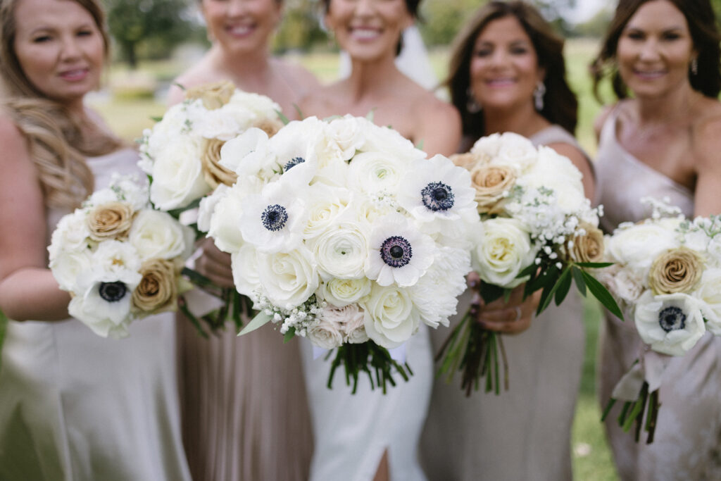 Five women in light-colored dresses hold bouquets of white and beige flowers with greenery, smiling outdoors on a sunny day. The focus is on the flowers in the foreground.
