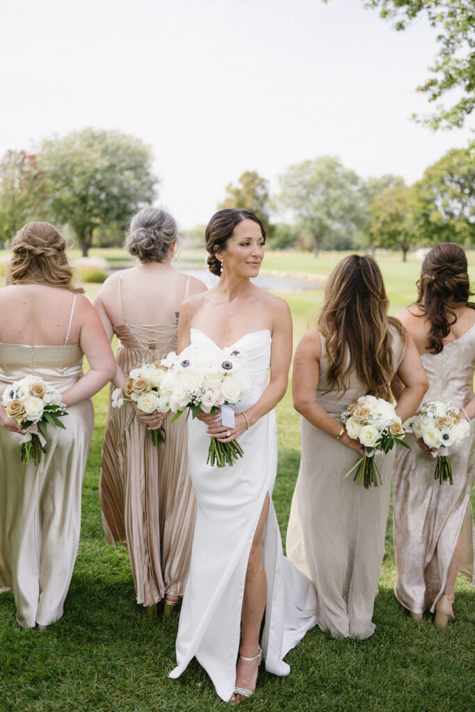 A bride in a white gown holds a bouquet and smiles while standing on grass with four bridesmaids in neutral dresses, all holding bouquets behind their backs. Trees and greenery are visible in the background.