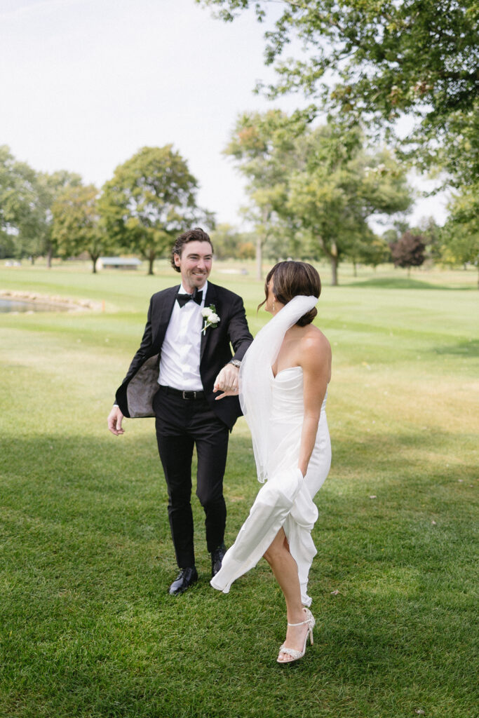 A bride and groom walk hand in hand on green grass outdoors, smiling at each other. The bride wears a white dress and heels, the groom wears a black tuxedo. Trees and blue sky are visible in the background.