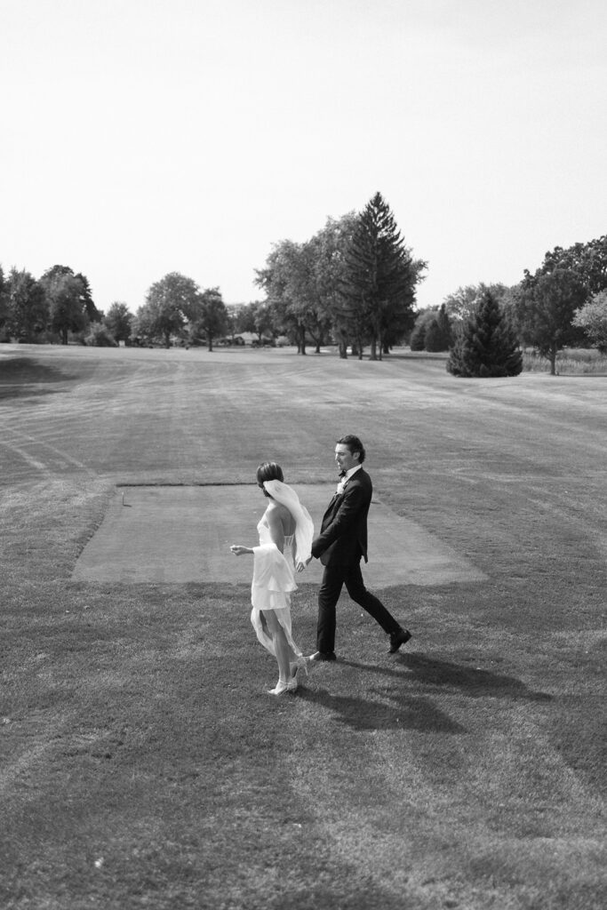 A woman in a short white dress and a man in a dark suit walk together across a grassy field on a sunny day, with trees in the background. The photo is in black and white.