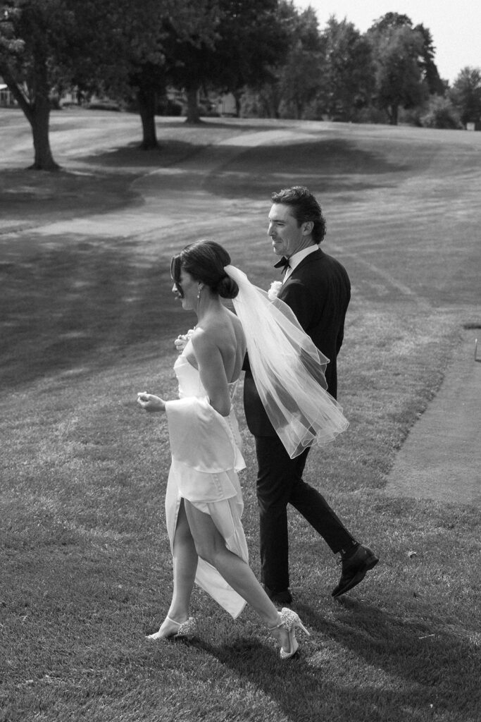 A bride in a strapless gown and veil walks on grass with a groom in a suit, holding hands outdoors on a sunny day, with trees and open lawn in the background. The image is in black and white.