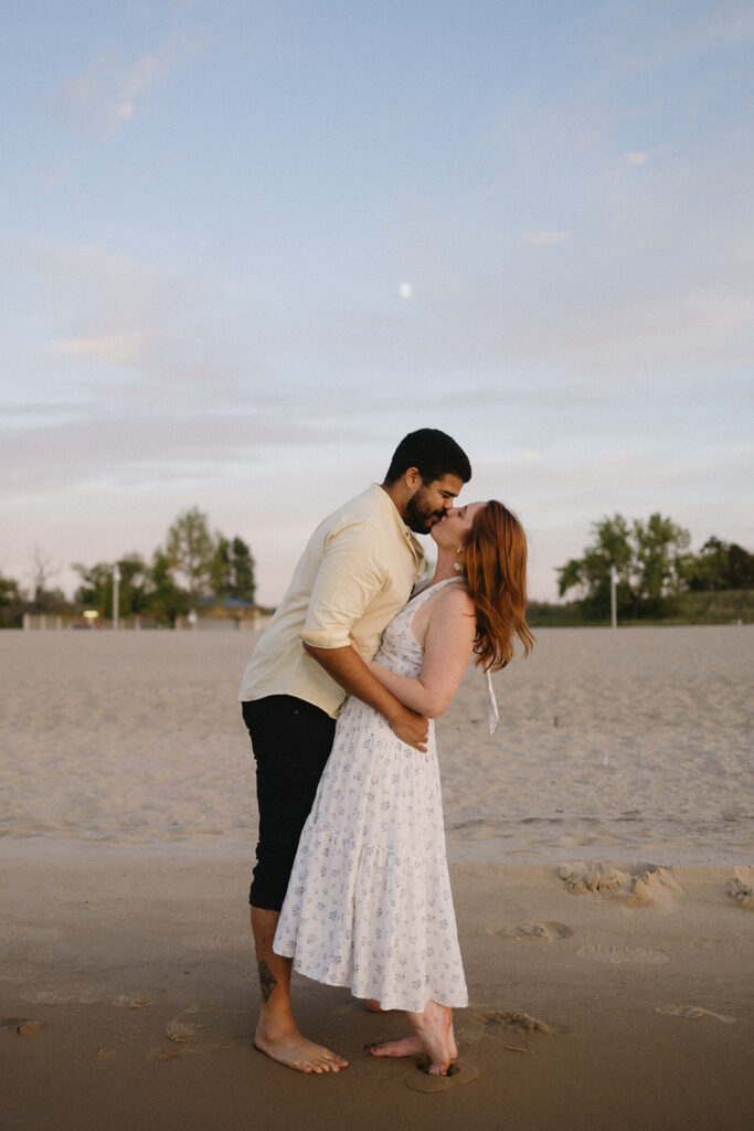 A couple stands barefoot on a sandy beach at sunset; the man, wearing a light shirt and dark pants, gently dips and kisses the woman, who is in a white dress. They appear happy and affectionate, surrounded by a tranquil scene.