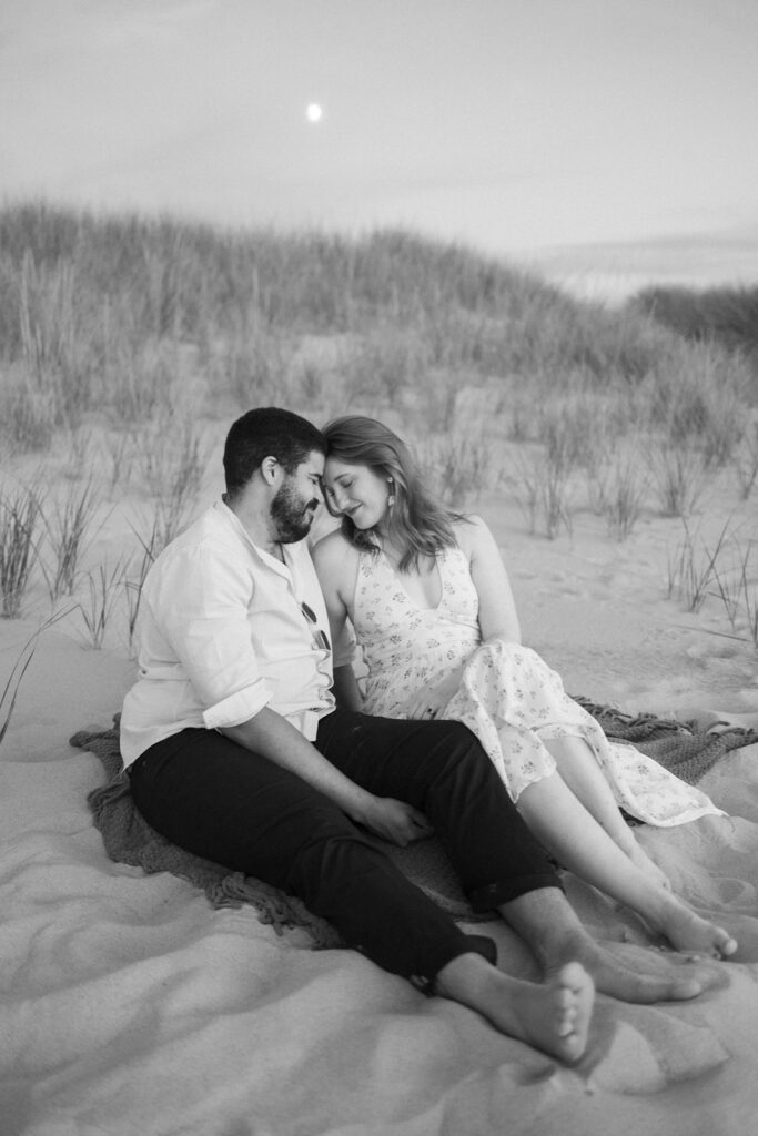A couple sits closely together on a blanket at the beach, smiling and touching foreheads. They are barefoot, surrounded by sand and tall grass, with the moon visible in the sky above. The image is in black and white.