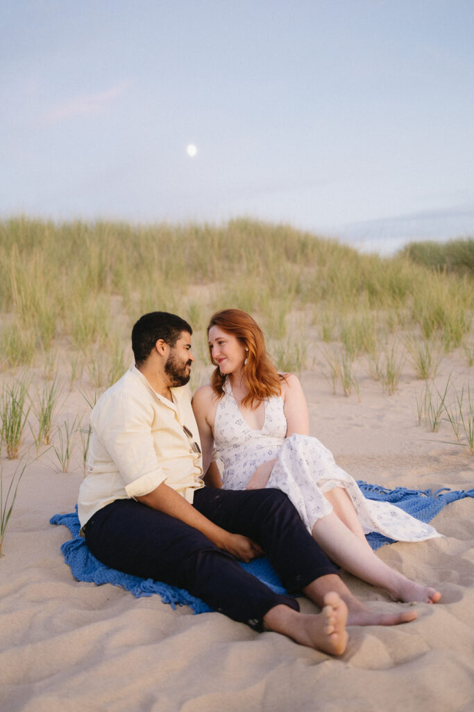A couple sits closely together on a blue blanket in sandy dunes, surrounded by grass. They gaze at each other, smiling affectionately, under a pastel evening sky with the moon visible above.