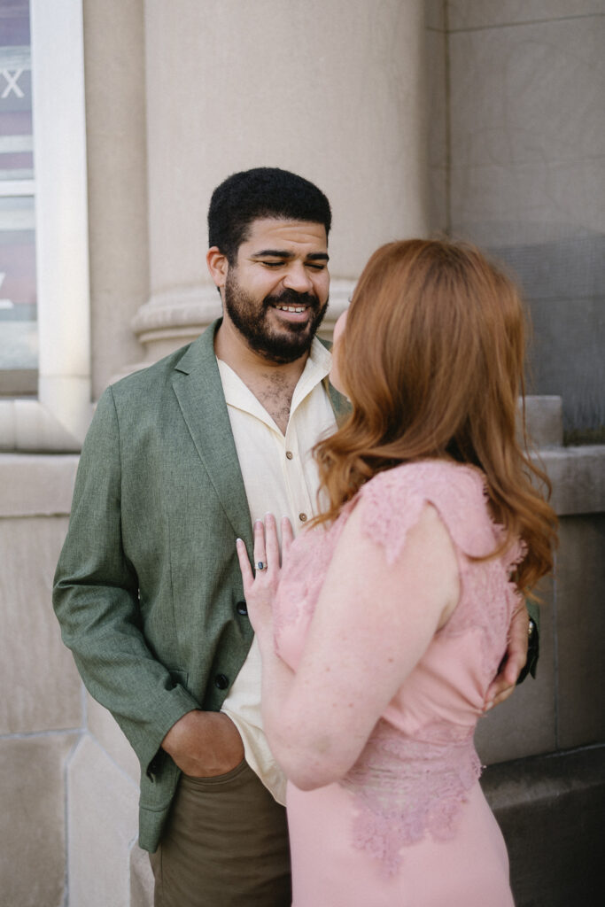 A man in a green blazer smiles at a woman in a pink lace dress as they stand close together outside a stone building. The woman has her hand gently resting on his chest.