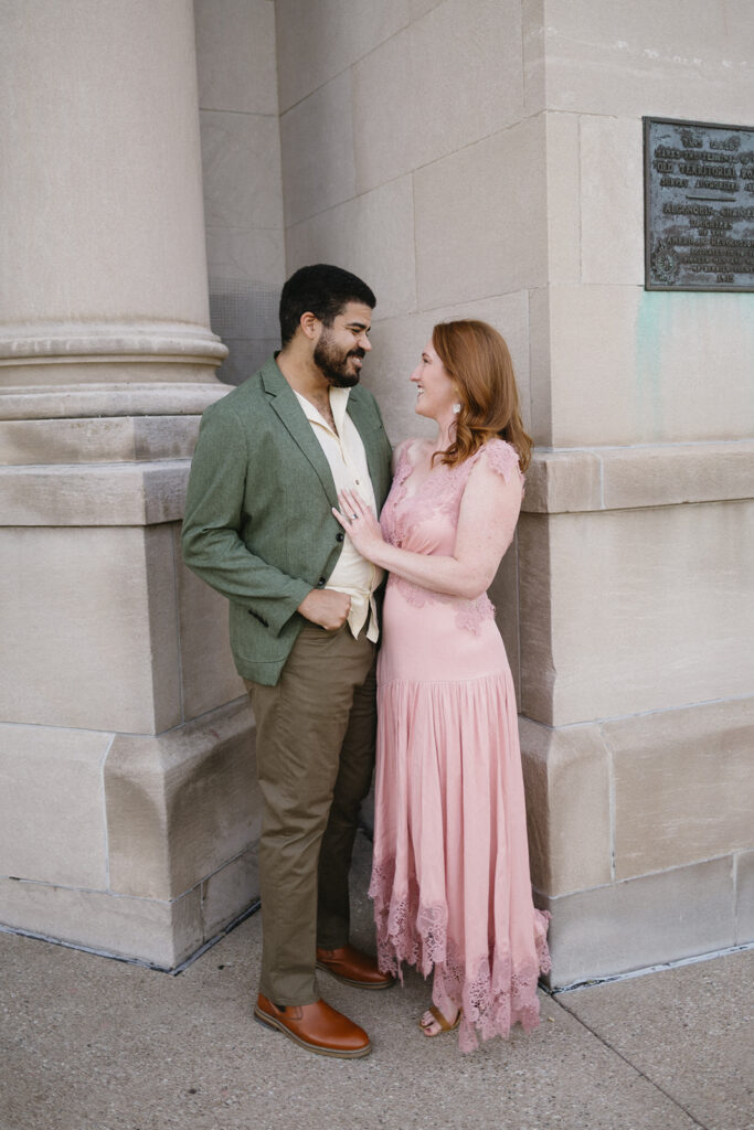 A man in a green jacket and khaki pants smiles at a woman in a long pink dress as they stand close together outside a stone building, looking at each other happily.