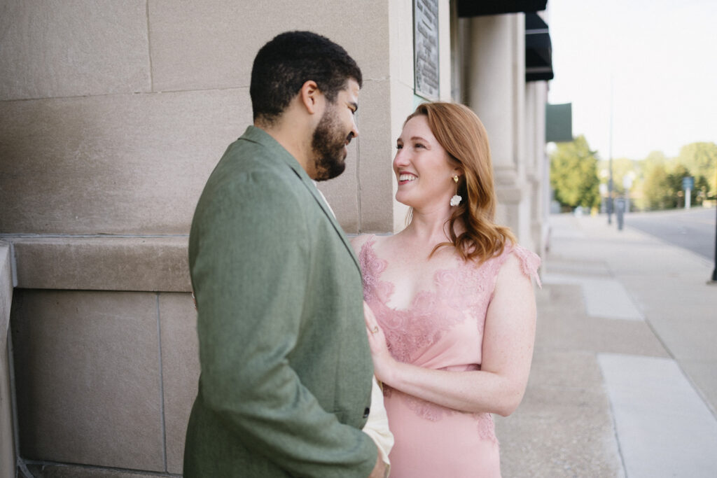 A man in a green suit and a woman in a pink lace dress stand close together, smiling warmly at each other beside a light stone building on a sunny day.