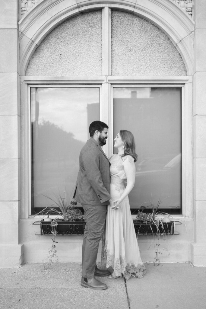 A couple stands facing each other, holding hands and smiling, in front of a large arched window with a stone frame. The image is in black and white.