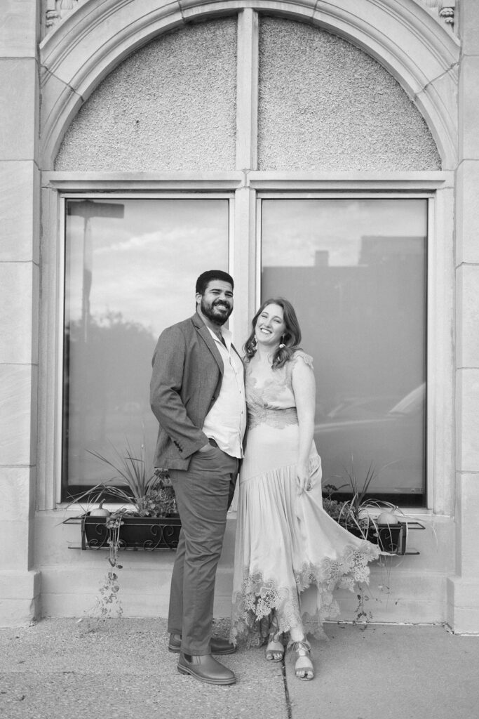 A smiling couple stands close together in front of a large arched window. The man wears a blazer and pants, and the woman wears a flowing dress. Both appear happy and relaxed. The photo is black and white.