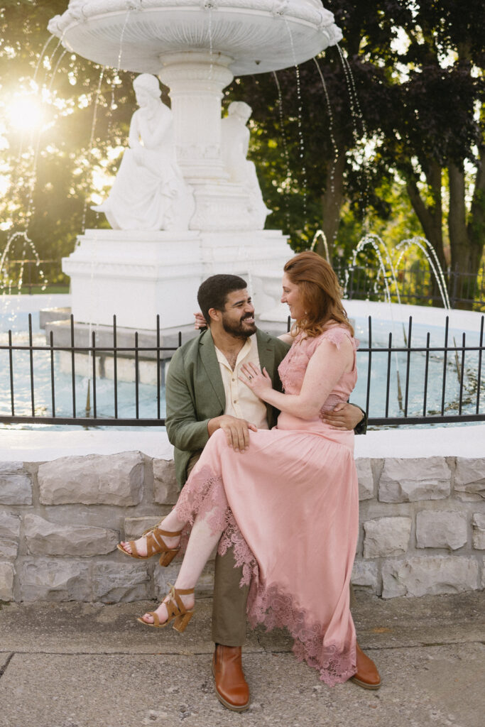 A couple sits closely together on a stone ledge in front of a white fountain. The man, in a green jacket, smiles at the woman, who wears a pink dress and heels. Sunlight streams through trees in the background.