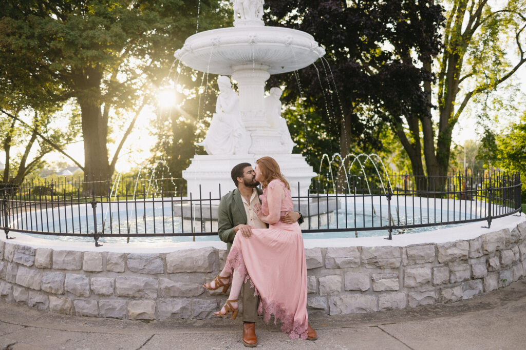 A couple poses affectionately in front of a white stone fountain in a park. The woman in a pink dress sits on the fountain’s edge, leaning into a man in a green suit as sunlight filters through the trees behind them.