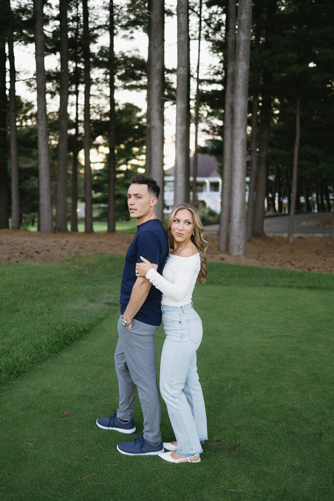 A man and woman stand back-to-back on grass in a wooded area. The woman, wearing a white top and jeans, holds the mans arm while he looks away. Tall trees and a house are visible in the background.