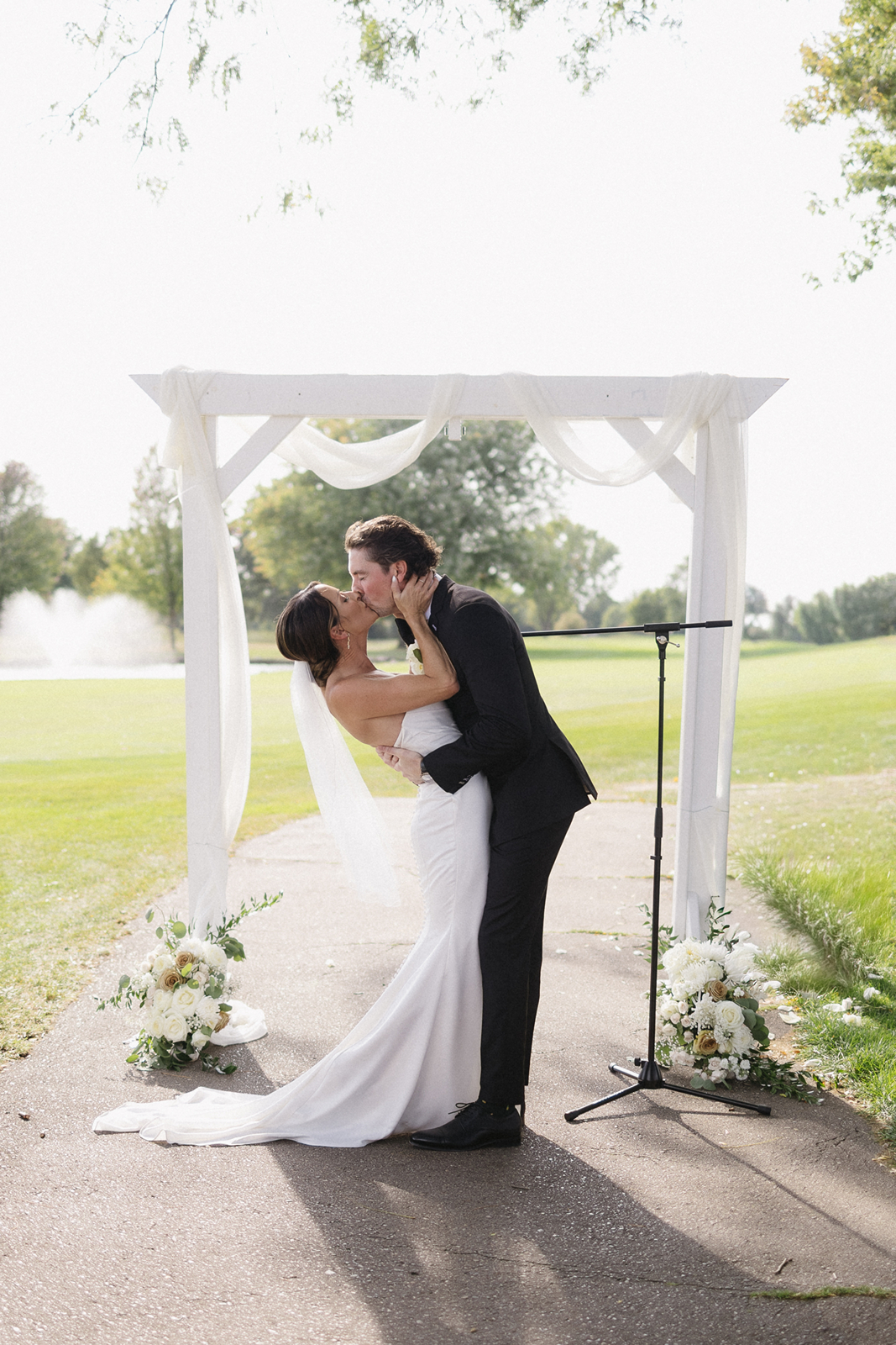 A bride and groom share a kiss under a white archway draped with fabric, surrounded by floral arrangements, outdoors on a sunny day with green grass and trees in the background.