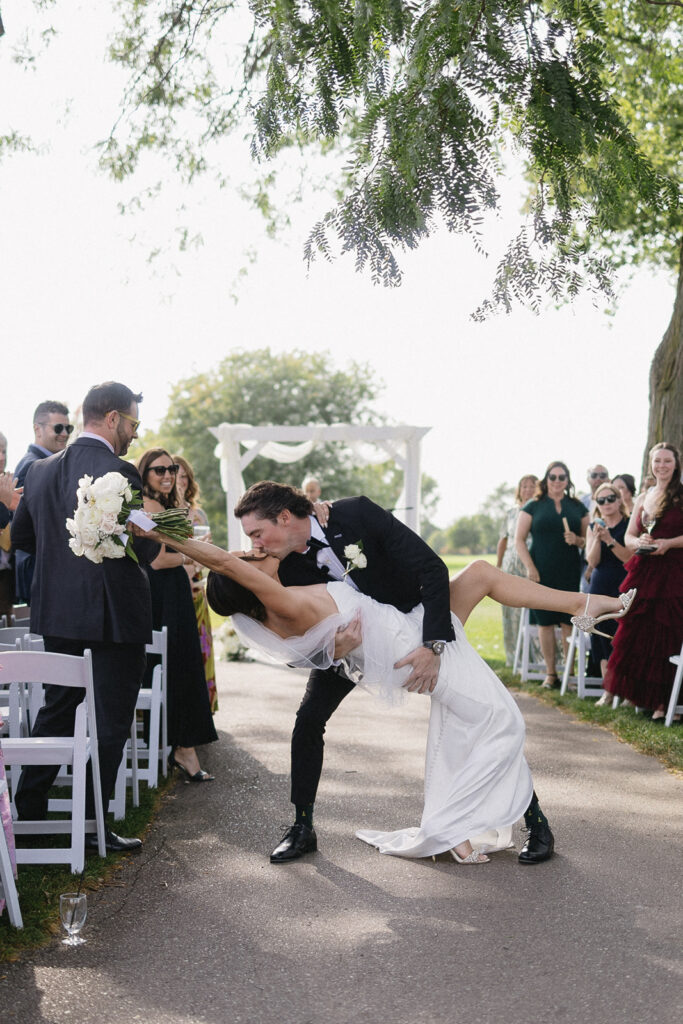 A groom in a black tuxedo dips his bride in a white dress for a kiss as they walk down the outdoor aisle, surrounded by applauding guests seated on white chairs.