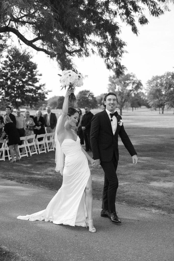 A bride and groom walk hand in hand outdoors. The bride, in a white dress, joyfully lifts her bouquet in the air. The groom wears a black tuxedo. Guests seated on white chairs watch in the background. The scene is cheerful and celebratory.
