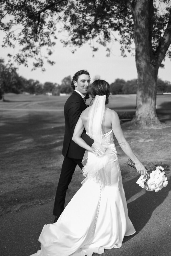 A bride and groom walk outside on a path, holding hands. The bride wears a long, fitted gown with a veil and holds a bouquet; the groom wears a suit. They look at each other, surrounded by trees and open grass.