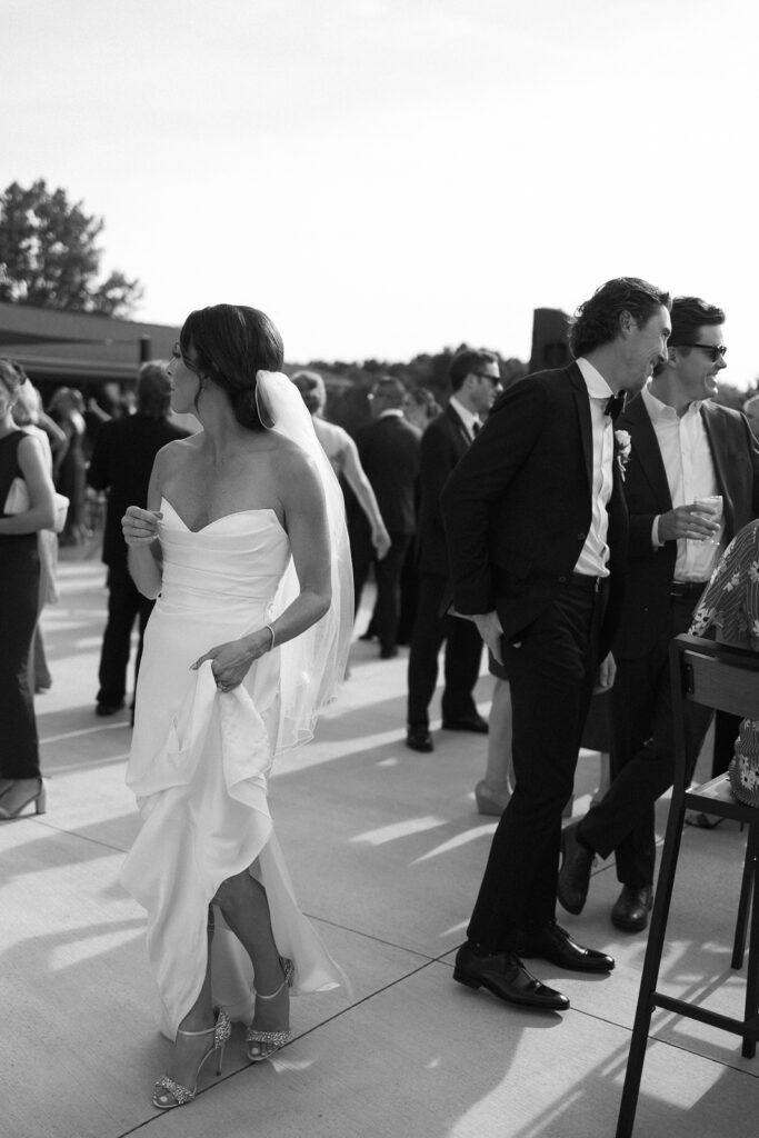 A bride in a strapless gown and veil walks outdoors at a wedding reception, holding her dress. Guests in formal attire stand and mingle in the background. The photo is in black and white.