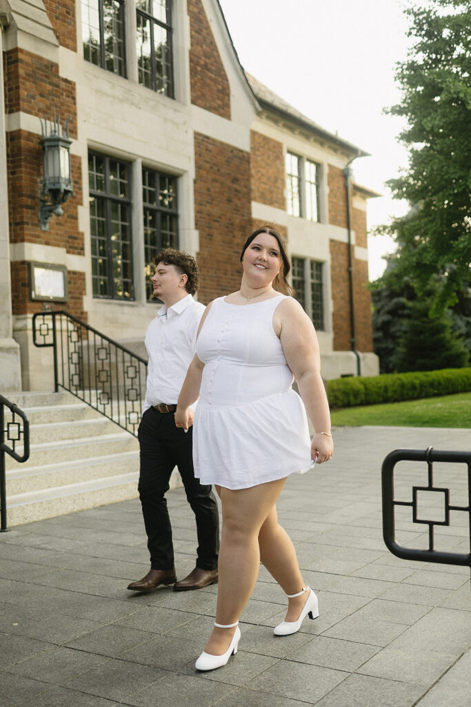 A woman in a white dress and heels smiles while walking in front of a brick building, with a man in a white shirt and black pants walking slightly behind her. The scene is outdoors on a paved walkway.